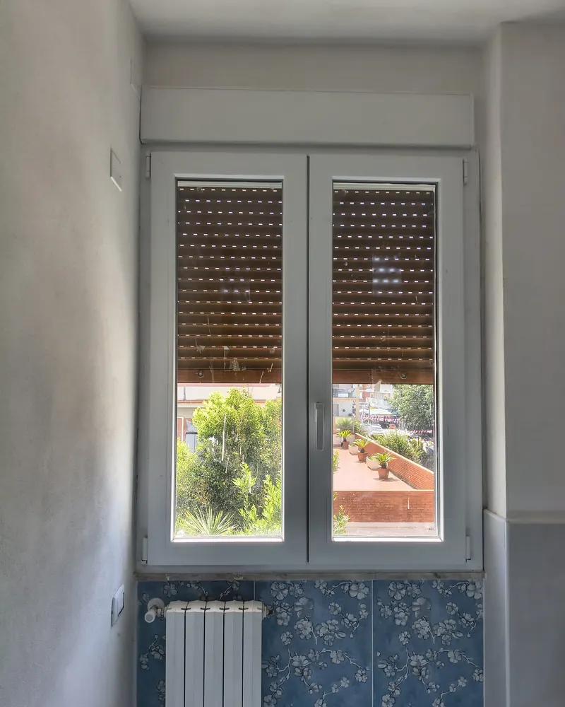 Finestra bianca a due ante in PVC vista dall’interno di un bagno, con tapparella e radiatore sotto